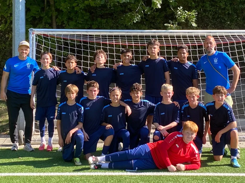 Die U16-Fußballmannschaft der Adolf-Reichwein-Schule steht gemeinsam mit ihren beiden Trainern vor einem Fußballtor. Die Spieler tragen dunkelblaue Trikots und Shorts. Im Vordergrund liegt der Torwart in einem roten Trikot auf dem Kunstrasen. Links steht Sportlehrkraft und Trainer Herr Strasser, rechts Sportlehrkraft und Trainer Herr Schulz. Die Mannschaft und ihre Trainer schauen in die Kamera. Im Hintergrund sind das Fußballtor und Bäume zu sehen.
