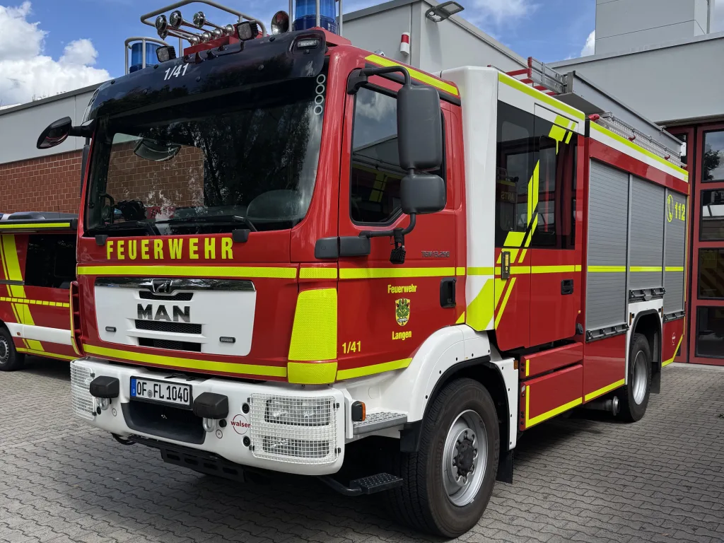 Ein rotes Löschfahrzeug der Feuerwehr Langen steht auf dem Hof der Feuerwache. Das Einsatzfahrzeug ist von vorne und von der rechten Seite zu sehen. Auf der Fahrzeugfront steht „FEUERWEHR“, an der Seite sind das Wappen und der Schriftzug „Feuerwehr Langen“ angebracht. Das Fahrzeug verfügt über eine große Mannschaftskabine, seitliche Geräteräume mit geschlossenen Rollläden und eine auffällige gelb-grüne reflektierende Beklebung. Im Hintergrund sind Teile der Feuerwache und ein blauer Himmel mit Wolken zu sehen.