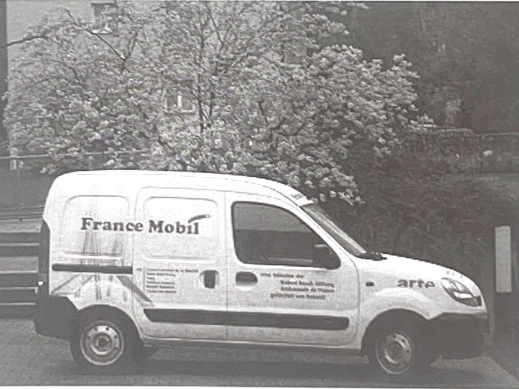 Auf dem Foto steht ein weißer Kleintransporter mit dem Schriftzug „FranceMobil“ auf der Seite auf dem Schulhof der Adolf‑Reichwein‑Schule. Das Fahrzeug ist vorne und seitlich zu sehen, es hat Fenster und wirkt wie ein klassischer Lieferwagen. Im Hintergrund sind Schulgebäude und Bäume erkennbar.