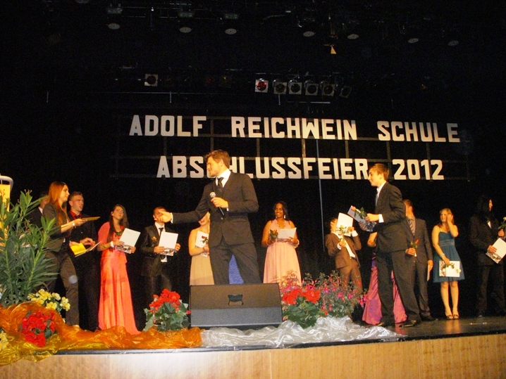 Schüler stehen auf einer Bühne bei einer Abschlussfeier und erhalten Urkunden.