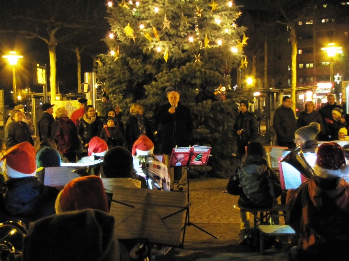 Menschen stehen um einen beleuchteten Weihnachtsbaum, während ein kleines Orchester spielt.