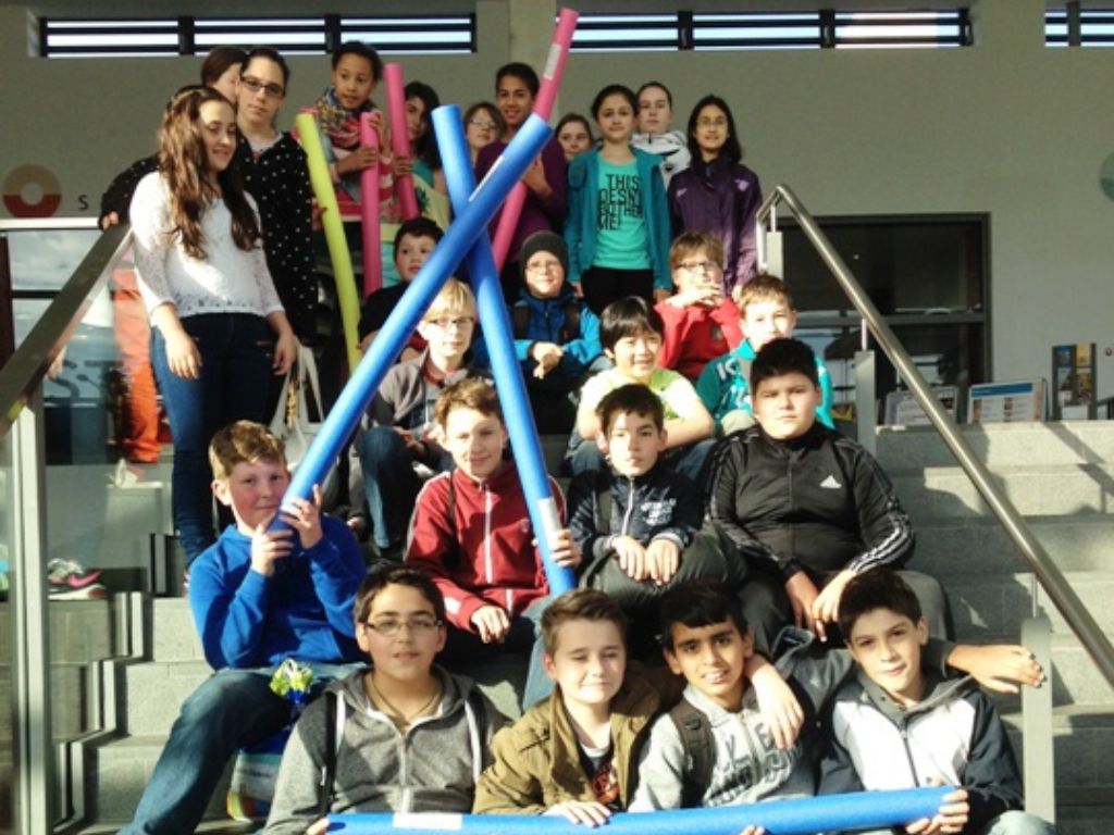 Auf dem Bild ist eine Gruppe von etwa 20 Schülern einer 5. Klasse zu sehen, die im Eingangsbereich des Langener Hallenbads auf einer Treppe sitzen und stehen. Die Treppe hat links und rechts Geländer aus Metall. Einige Schüler halten bunte Schaumstoffstäbe in Blau, Pink und Gelb in den Händen. Die Kinder tragen unterschiedliche Alltagskleidung, darunter Jacken, Pullover und Brillen. Im Hintergrund sind Fenster und Wände des Hallenbad-Eingangsbereichs sichtbar. Die Gruppe schaut größtenteils zur Kamera und wirkt freundlich und entspannt.