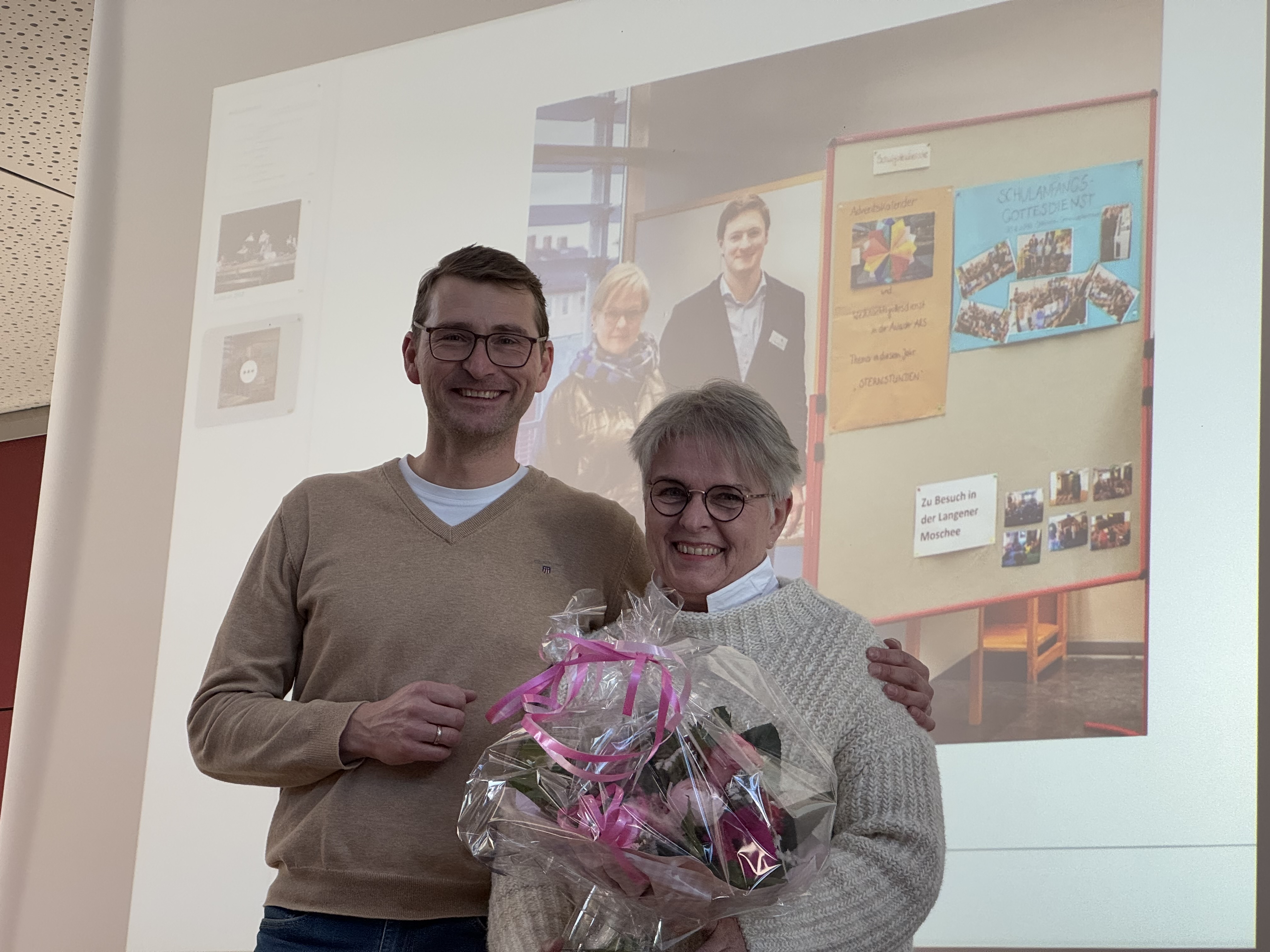 Auf dem Foto stehen zwei Erwachsene im hellen Innenraum der Adolf‑Reichwein‑Schule vor einer großen Projektionsfläche. Links steht der Schulleiter, ein Mann mit kurzen Haaren und Brille, in einem hellen Pullover über einem weißen T‑Shirt. Rechts steht eine Frau mit kurzen, hellgrauen Haaren und runder Brille; sie hält einen Blumenstrauß mit rosa Geschenkband und lächelt zur Kamera. Die Szene zeigt den Abschied der Kollegin in den Ruhestand. Im Hintergrund sind auf der Projektionsfläche unscharf ein Foto von mehreren Personen und bunte Pinnwände zu erkennen.
