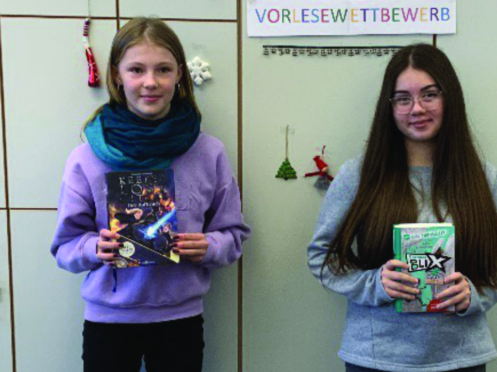 Auf dem Foto stehen zwei Schülerinnen nebeneinander vor einer hellen Wand oder Schrankreihe. Über ihnen hängt ein Schild mit dem Wort „VORLESEWETTBEWERB“, das ankündigt, worum es bei der Szene geht. Die Schülerin links trägt einen hellen Pullover und einen dunkelgrünen Schal, sie hält ein Buch mit farbigem Cover in den Händen. Die Schülerin rechts hat lange, offene Haare und eine Brille; auch sie hält ein Buch mit einem grünen Einband. Beide schauen leicht zur Kamera und stehen dicht beieinander. Im Hintergrund sind an der Wand kleine Deko‑Elemente zu erkennen.