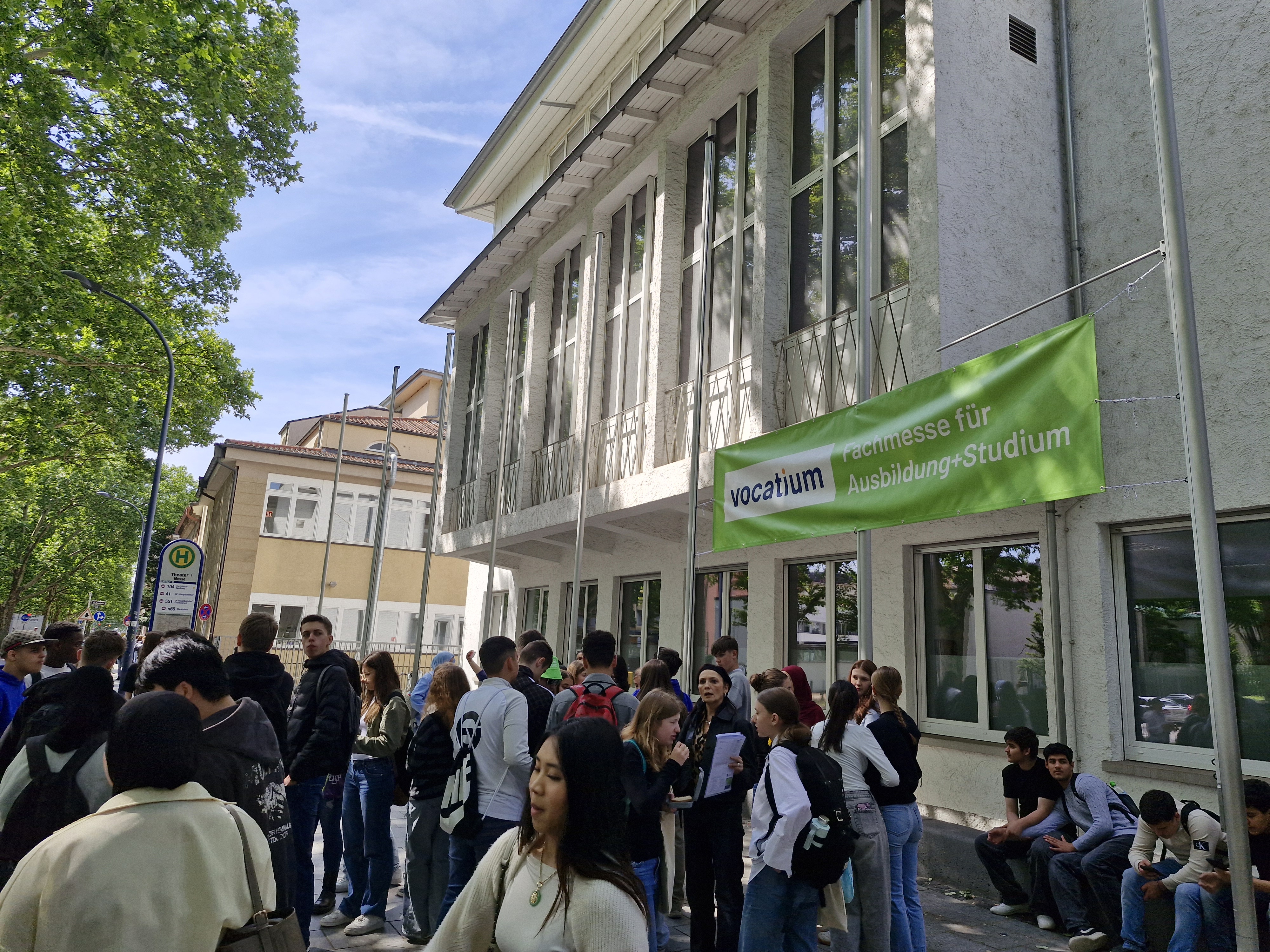 Das Bild zeigt einen Computerbildschirm mit einem Artikel über eine Berufsmesse.    In der Mitte ist ein Foto von vielen Schülerinnen und Schülern, die draußen vor einem Gebäude stehen und sich informieren. Man sieht Logos und Banner der Messe „vocatium“.    Darunter steht ein Text darüber, dass die Jugendlichen die Messe besucht haben, um sich über Berufe und Studienmöglichkeiten zu informieren. Rechts ist ein kleines Porträt von einem Ansprechpartner.